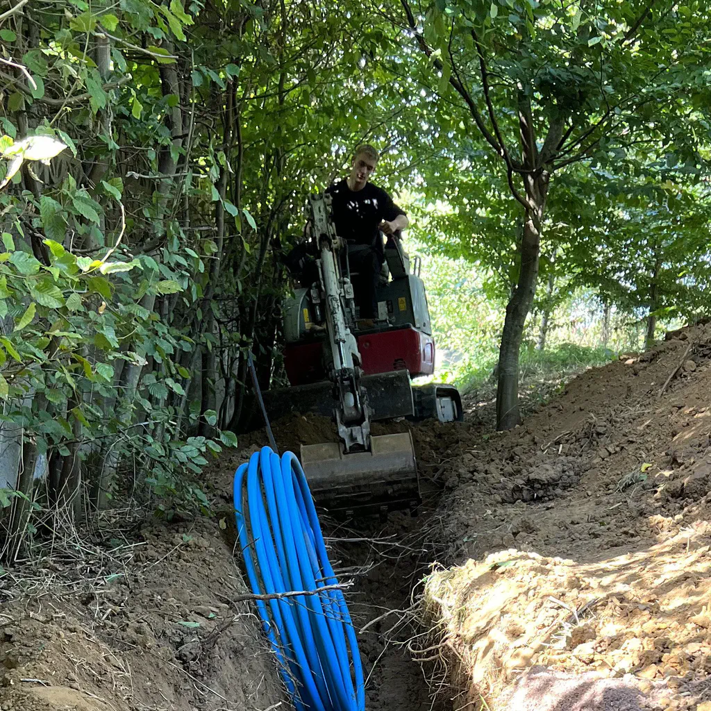 A CTC operator digging a service trench through woodland, with a coil of blue water main ready to lay